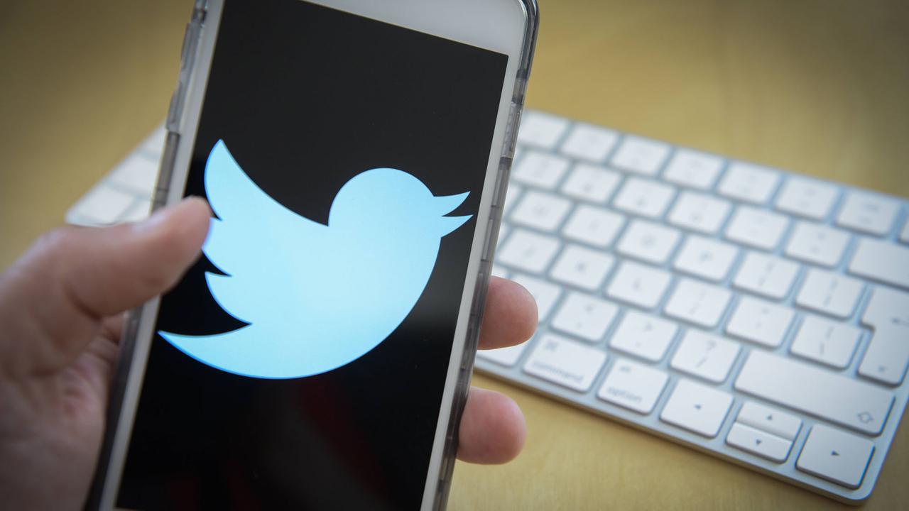 Das Twitter-Logo auf einem Mobiltelefon, gehalten von einer Hand, im Hintergrund eine Computertastatur