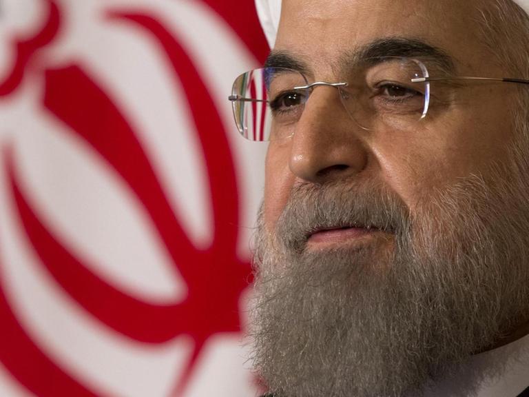 Rohani trägt einen weißen Turban und Brille. Hinter ihm sieht man einen teil der iranischen Nationalflagge.
