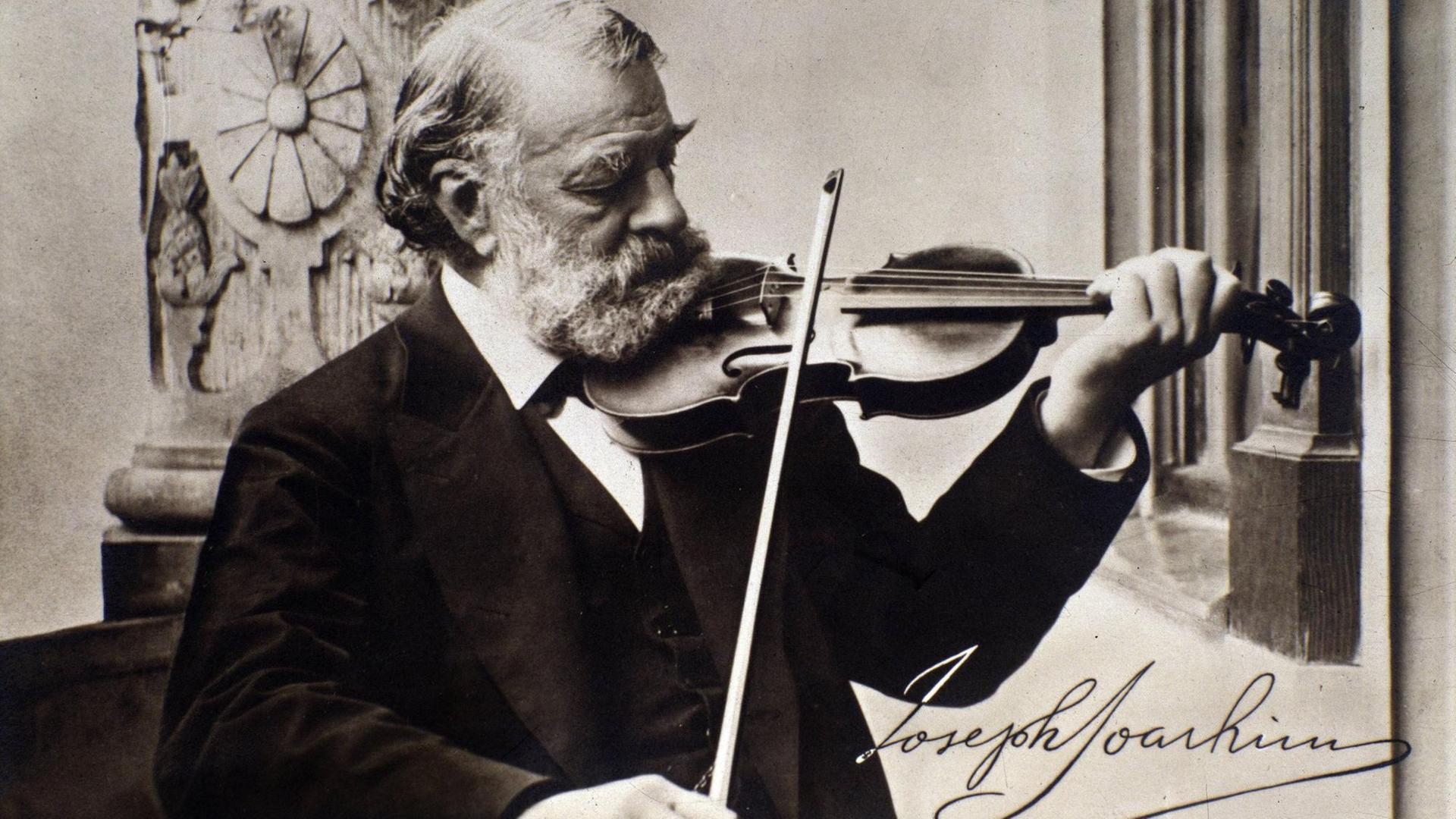 Schwarz-weiß Foto, aufgenommen um 1900, mit dem spielenden Geiger Joseph Joachim, er trägt einen scharzen Anzug und schaut auf das Griffbrett, er trägt einen weißen Vollbart und hat weiße Haare, unten rechts ist seine Unterschrift verzeichnet