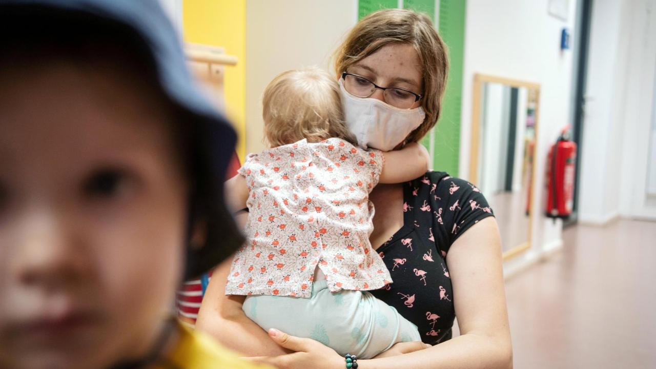 Eine Erzieherin mit Mundschutz, hält einen ihrer Schützlinge in einer Kindertagesstätte im Arm. 