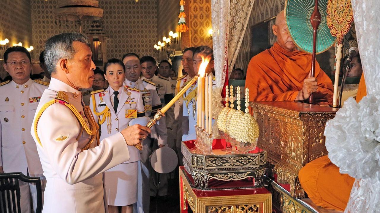 Vajiralongkorn steht an einer Art Schrein, auf dem ein buddhistischer Mönch sitzt. Vajiralongkorn zündet mit einer Kerze mehrere weitere an. Dahinter Hofstaat.  