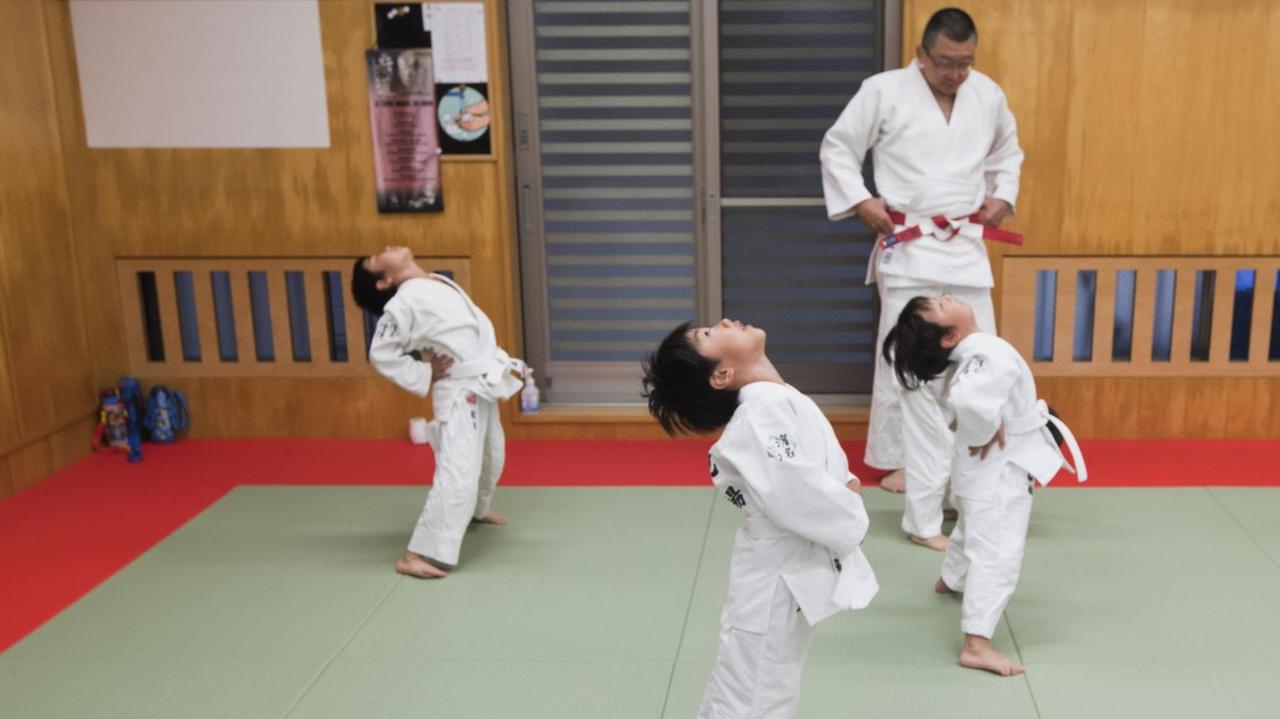 Judotraining: Kinder stehen in einer Sporthalle und dehnen ihren Rücken.
