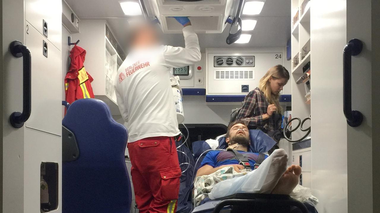 Pussy-Riot-Mitglied Pjotr Wersilow liegt bei seiner Ankunft in Berlin auf einer Liege im Krankenwagen. Ein Pfleger der Feuerwehr versorgt ihn, im Hintergrund eine Freundin.  