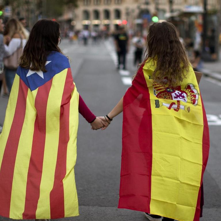 Zwei junge Frauen gehen nebeneinander. Ein von ihnen trägt eine Spanien-Flagge als Umhang, die andere eine Flagge von Katalonien.