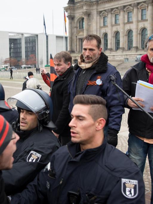 Vier Reichsbürger stehen hinter einem Mikrofoständer, einer von ihnen hält das Mikro. Drei Polizisten stehen davor, sie riegeln die Kundgebung gegen Gegendemonstranten ab.