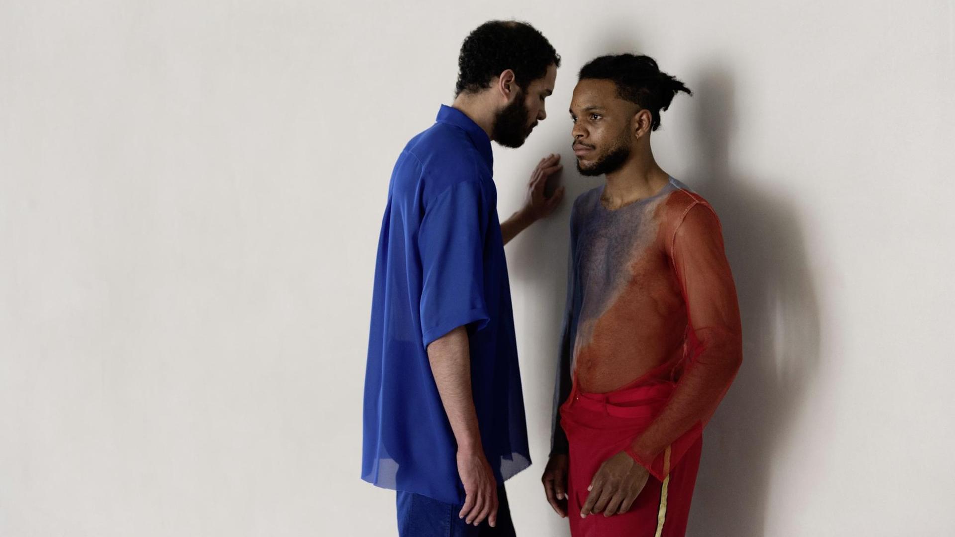 Ein Mann, in einem blauen Hemd, steht neben einem weiteren Mann, in einem halb transparenten roten Shit, der sich gegen die Wand lehnt.
