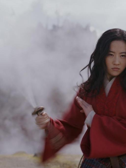 Schauspielerin Yifei Liu steht in der Rolle der Mulan in einer nebligen Landschaft. Sie nimmt eine Kämpferpose ein.