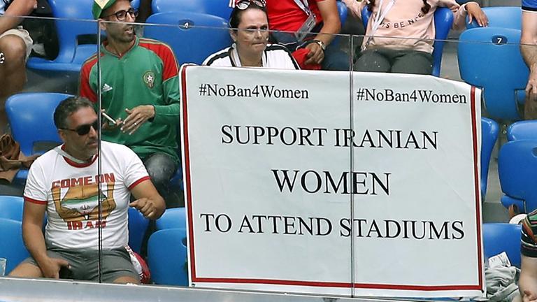 Das Bild zeigt eine Szene am Rande der Fußball-WM-Vorrundenbegegnung zwischen Marokko und dem Iran im Sankt-Petersburg-Stadion. Ein Poster zur Unterstützung von iranischen Frauen zum Besuch in Fußball-Stadien hängt an der Tribüne.