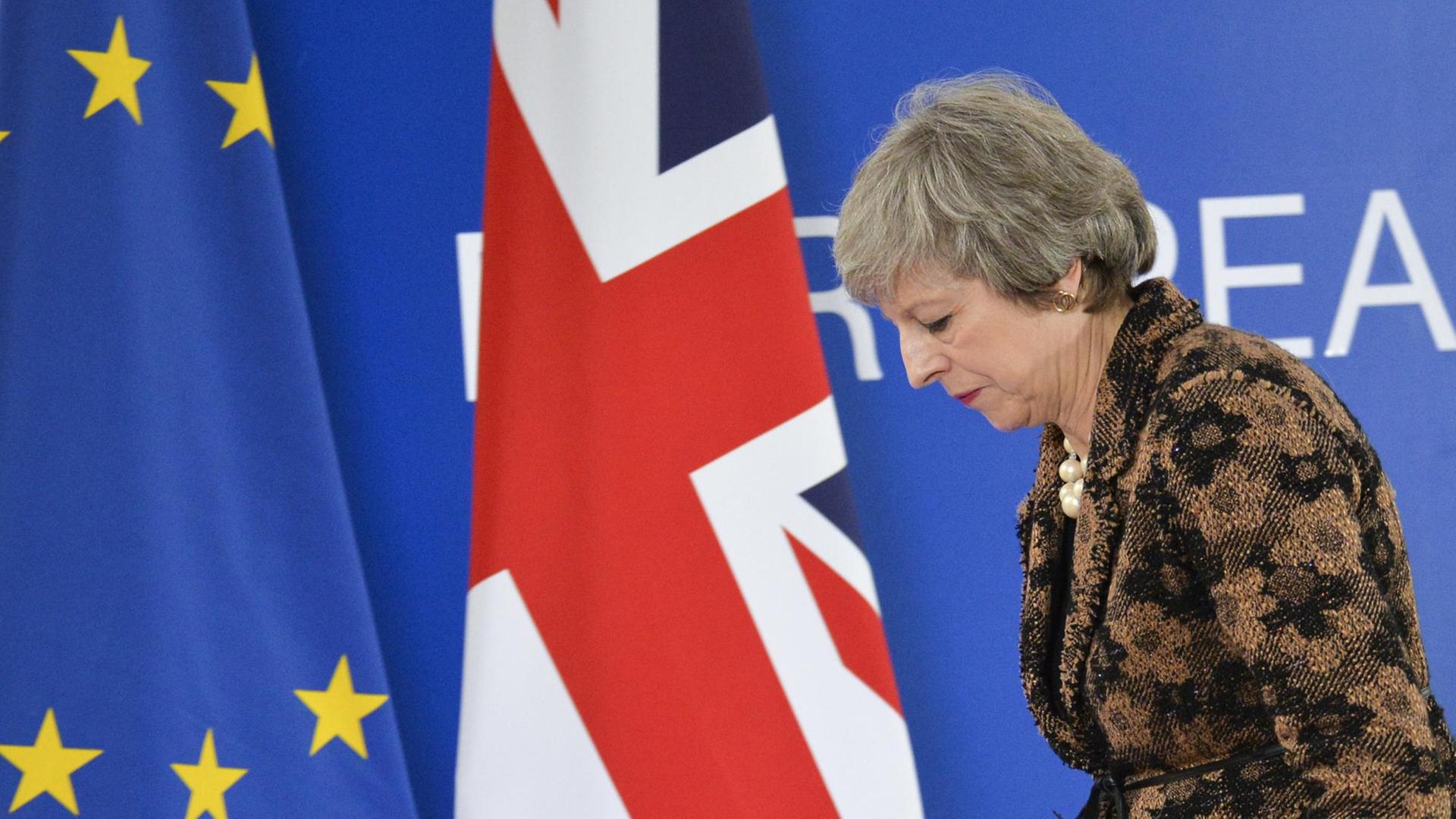 May läuft gebeugt an einer EU- und einer Gr0ßbritannien-Flagge vorbei.