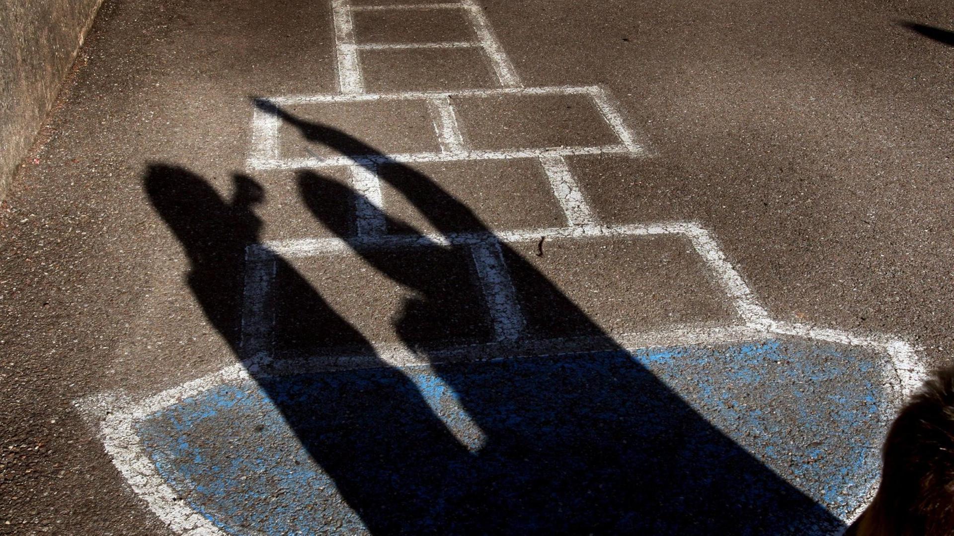 Schatten zweier Kinder auf einem Schulhof. Auf dem Boden ist ein Hüpfspiel mit Kreide aufgezeichnet.