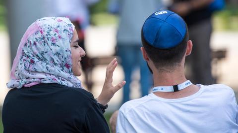 Eine junge Muslima mit Kopftuch und ein junger jüdischer Mann mit Kippa unterhalten sich miteinander. Sie sitzen auf einer Bank, vom Betrachter abgewandt.