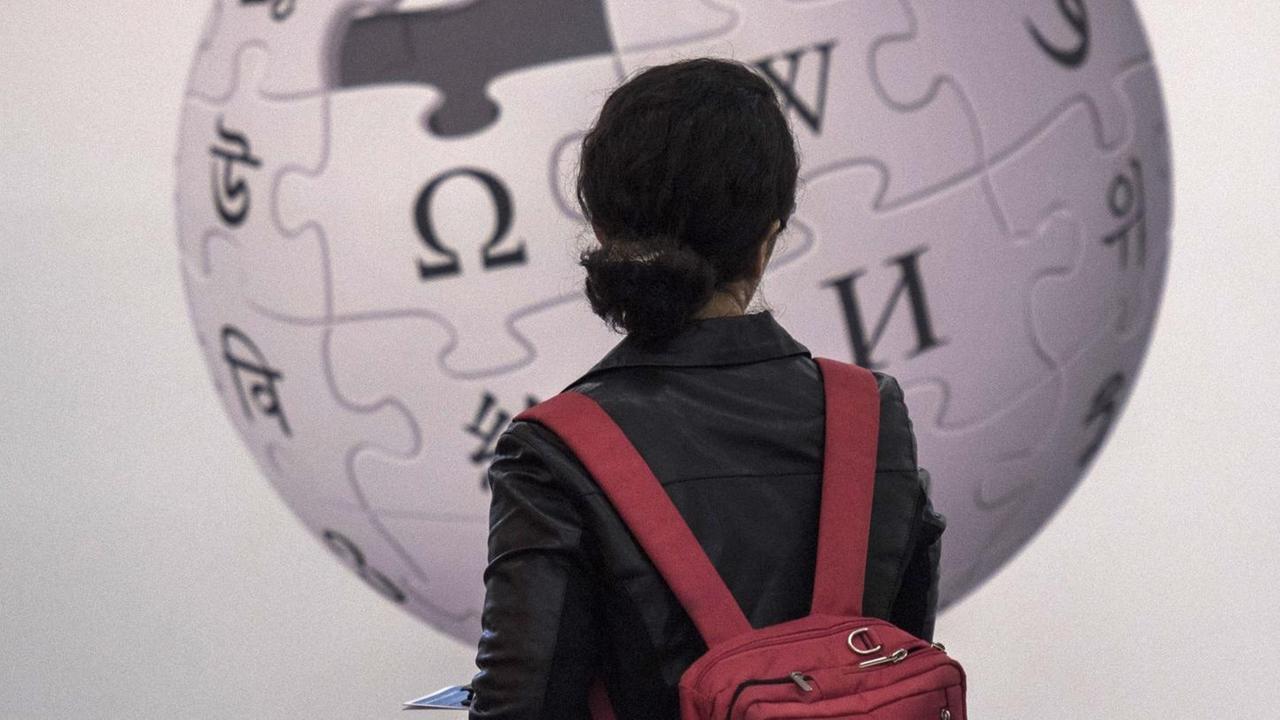 Eine Frau steht vor einer Wand und betrachtet das darauf angebrachte Logo der Wikipedia.