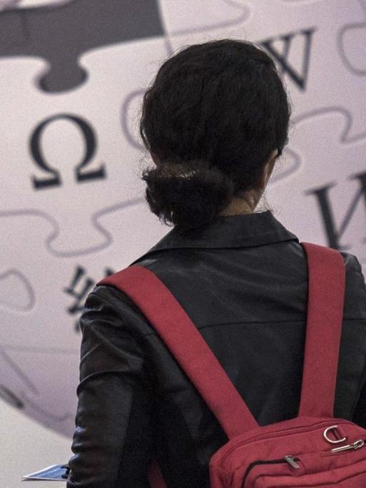 Eine Frau steht vor einer Wand und betrachtet das darauf angebrachte Logo der Wikipedia.