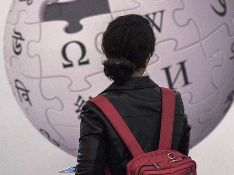 Eine Frau steht vor einer Wand und betrachtet das darauf angebrachte Logo der Wikipedia.
