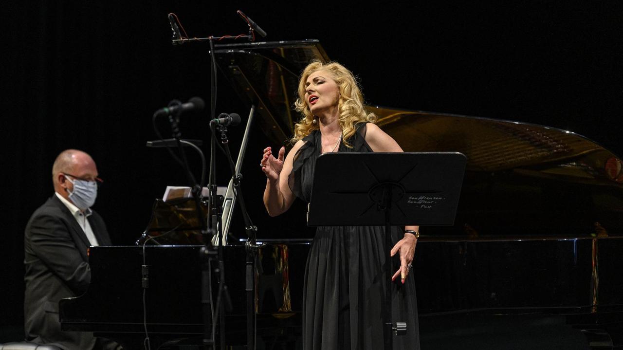 Eine blonde Sängerin in schwarzem Abendkleid steht neben einem Flügel samt Pianisten.