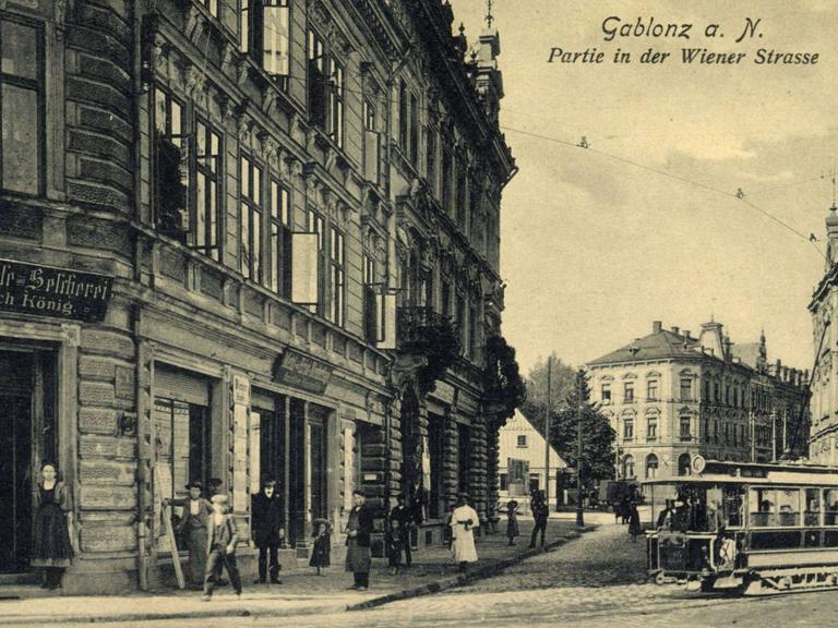 Historische Stadtansicht von Gablonz an der Neiße: Stadthäuser säumen eine Straße mit Kopfsteinpflaster, über die eine Straßenbahn fährt.