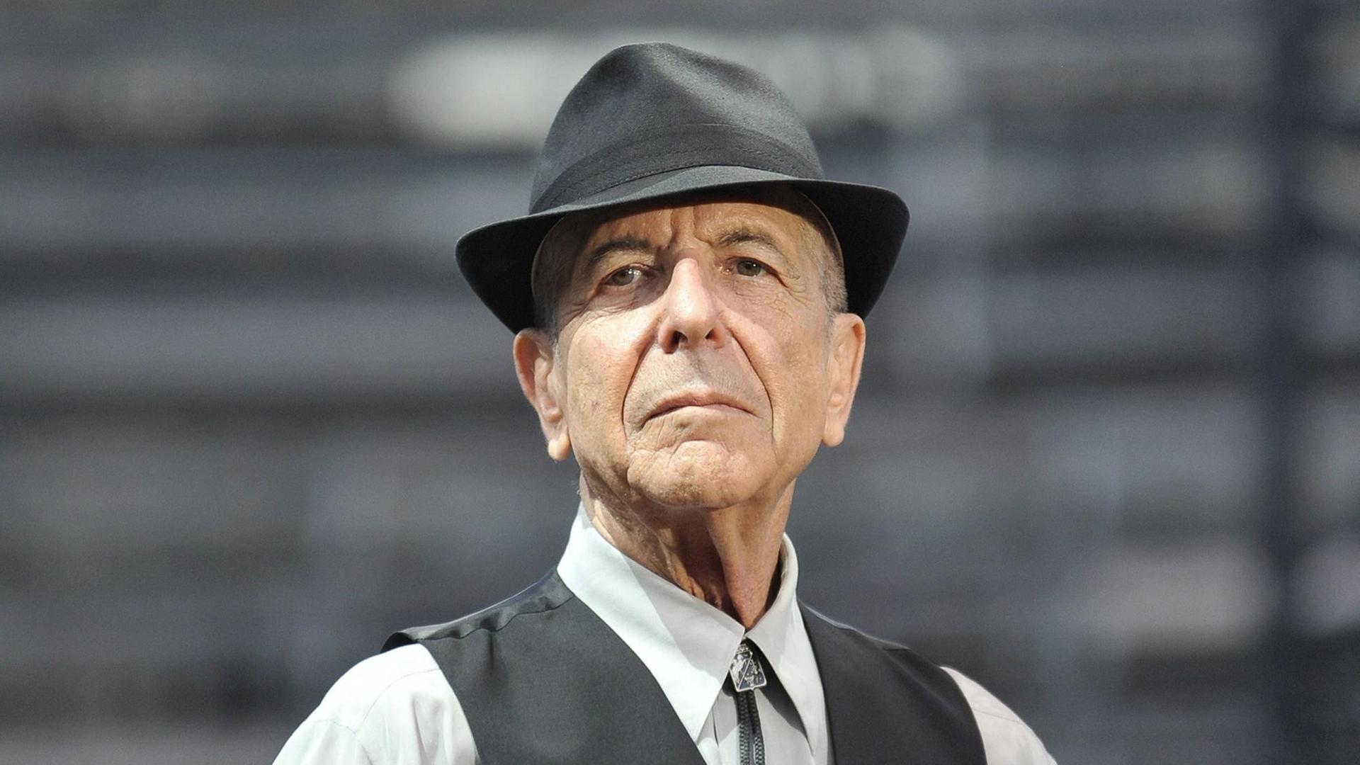 Leonard Cohen trägt ein helles Hemd, darüber eine dunkle Weste, außerdem einen Hut und schaut in die Kamera.