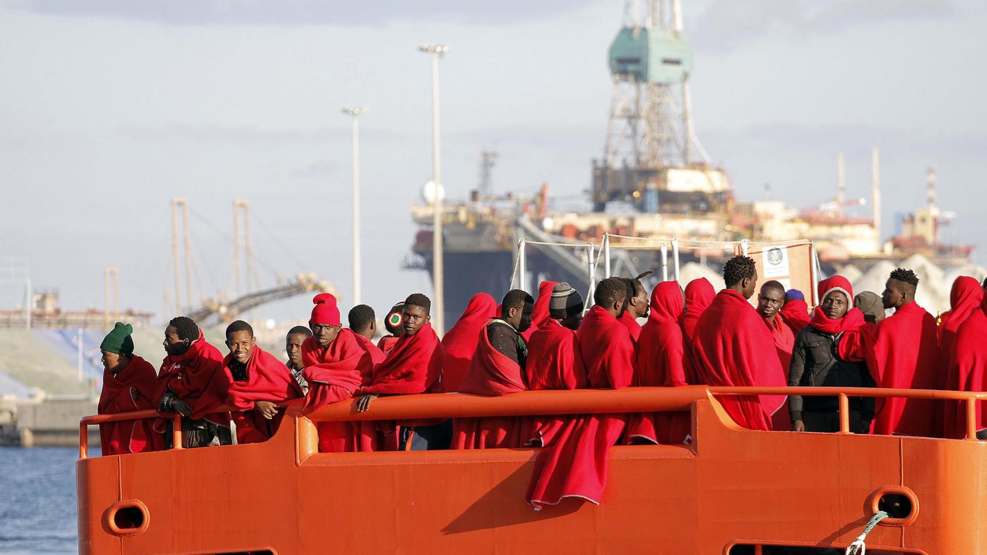 Gerettete Flüchtlinge aus der Subsahara stehen auf einem Boot der spanischen Küstenwache, eingewickelt in rote Decken.