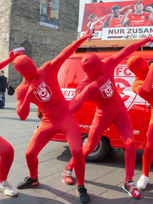 Vier Menschen in Ganzkörperkostümen, die auch das Gesicht verhüllen, stehen vor einem roten VW Bus, der mit Werbung des Halleschen FC bedruckt ist. Sie zeigen in den Himmel.