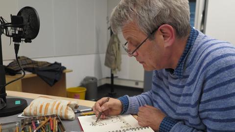 Der Illustrator Axel Scheffler zeichnet im Dlf-Studio einen "Grüffelo" in das Gästebuch der Sendung "Klassik-Pop-et cetera“, daneben liegt eine Box mit Buntstiften