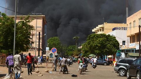Anschlag in Ouagadougou: Menschen stehen in einiger Entfernung zu dem Anschlagsort in der Hauptstadt von Burkina Faso. Man sieht dichte, schwarze Rauchwolken.