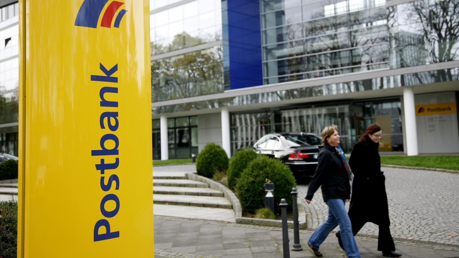 Blick auf den Eingangsbereichs; vor dem zwei Menschen vorbeilaufen. Im Vordergrund eine gelbe Stele mit dem Postbank-Schriftzug und dem Logo. Blick auf den Eingangsbereichs; vor dem zwei Menschen vorbeilaufen. Im Vordergrund eine gelbe Stele mit dem Postbank-Schriftzug und dem Logo.