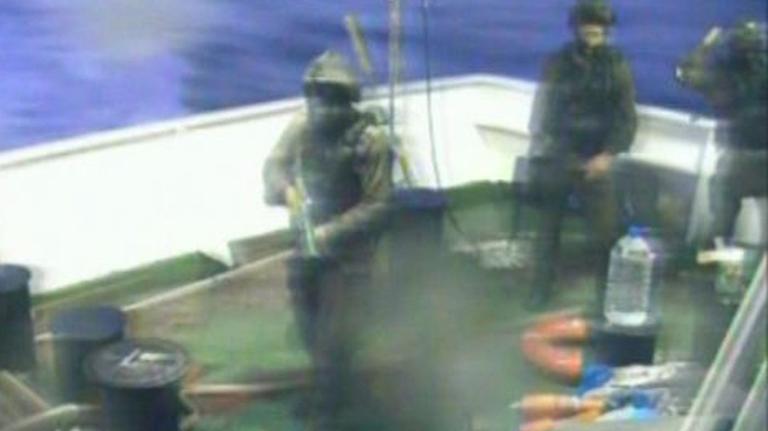 Man sieht drei bewaffnete Soldaten in Tarnuniformen und mit Helmen am Heck des Schiffes.