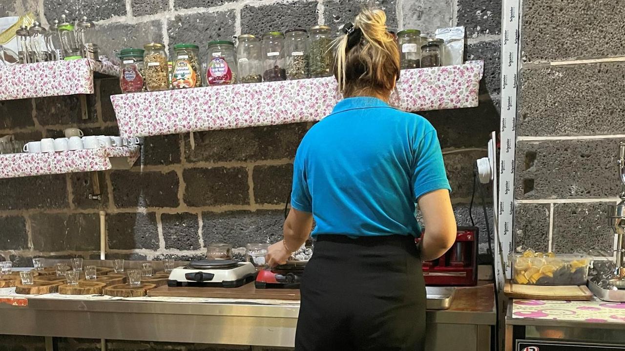 Eine junge Frau mit blondem Pferdeschwanz und T-Shirt steht in einer Küche an der Spüle und arbeitet.