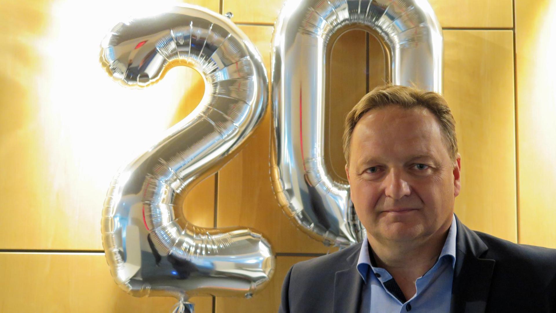 Jürgen Böhm, Vorsitzender Verband Deutscher Realschullehrer steht vor einer Holzwand. Im Hintergrund sind zwei silberne Luftballons zu sehen, welche die Zahl 20 bilden.
