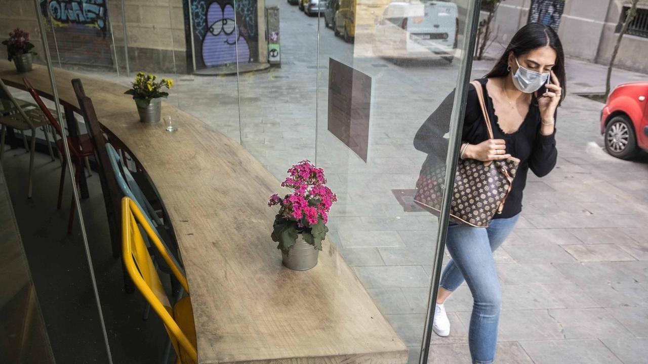 Eine junge Frau mit Coronamaske läuft in Barcelona an einem leeren Café vorbei.