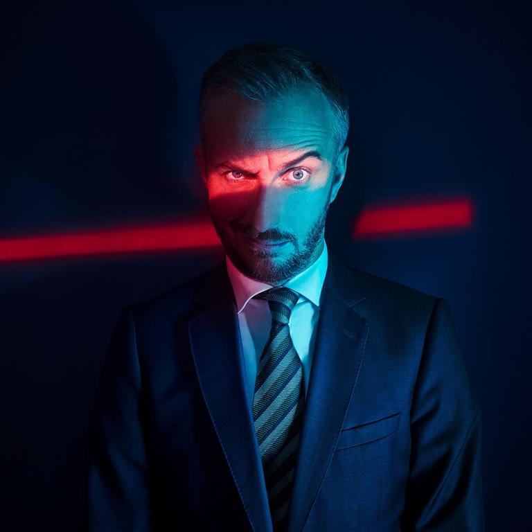 Jan Böhmermann blickt kritisch-spöttisch in die Kamera, eine Augenbraue hochgezogen. Er trägt Anzug und Krawatte und steht im Dunklen. Ein Spalt rotes Licht leuchtet auf seine Augenpartie und setzt diese so in den Fokus des Fotos.