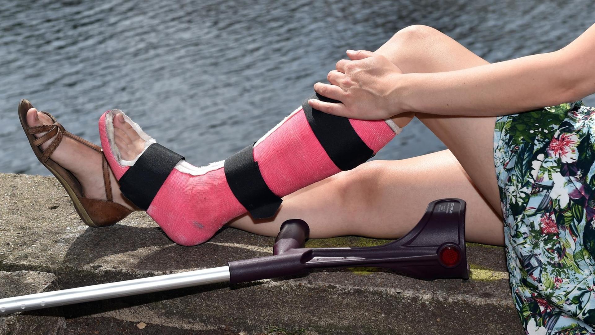 Eine junge Frau mit einem gebrochenen, rosa eingegipsten Bein sitzt am Ufer der Spree.
