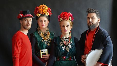 Zwei Männer und zwei Frauen in bunten Kleidern mit bunten Hüten aus Blumen stehen nebeneinander und blicken in die Kamera. Sie halten unterschiedliche Musikinstrumente in der Hand.