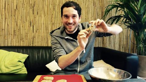 Farbfoto eines jungen Mannes, der stolz einen Burger in seiner Hand zeigt. Der besteht unter anderem aus Insekten.