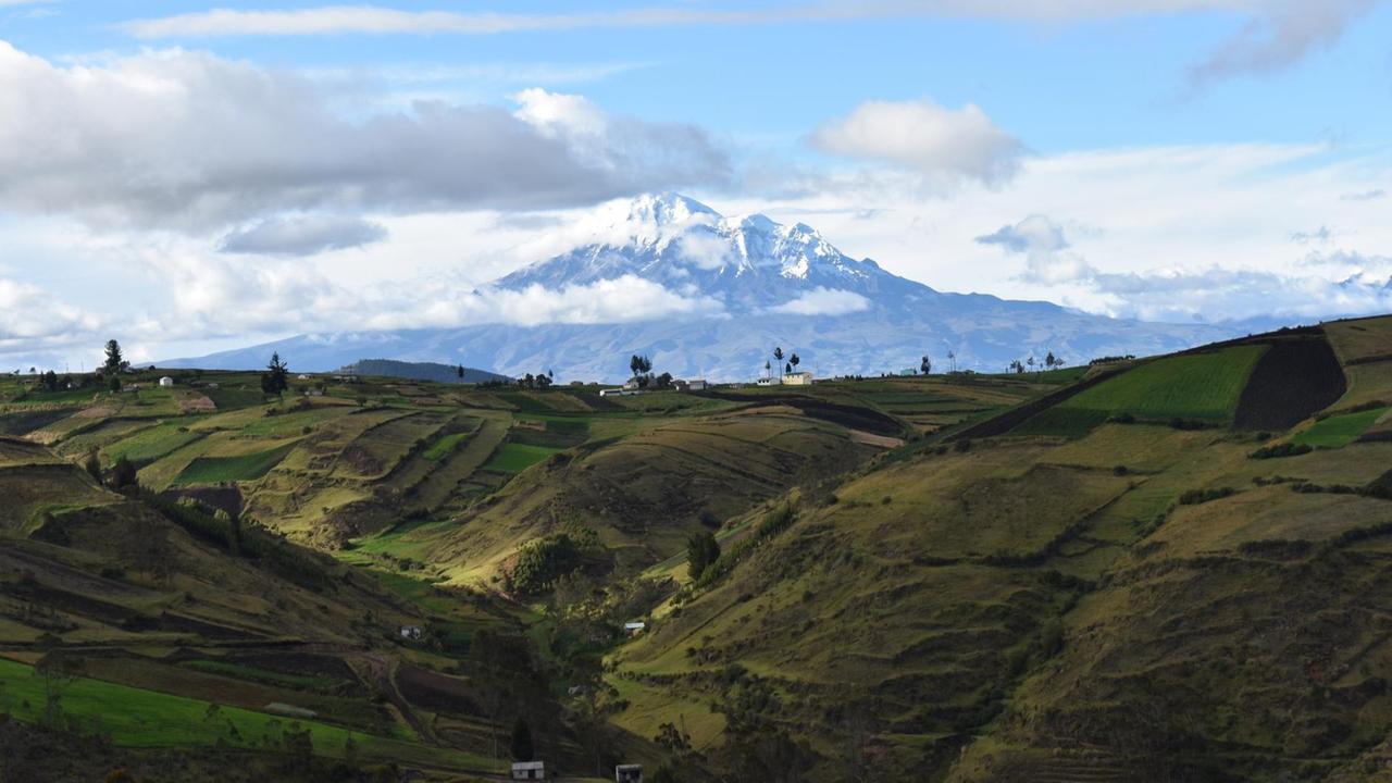 Panoramaaufnahme eines Vulkans mit verschneiter Kuppe vor einer grünen Hügellandschaft