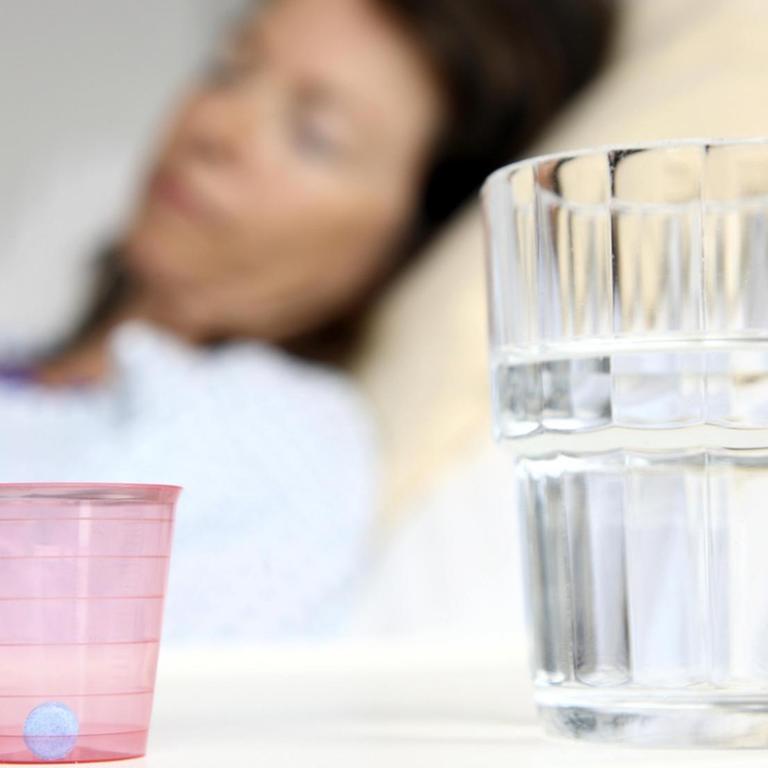 Eine Patientin liegt in einem Krankenbett, Medikament und Wasserglas auf dem Nachttisch im Bildvordergrund
