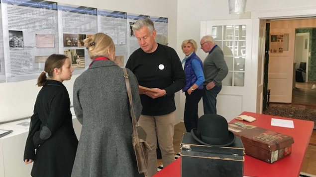 Im Ausstellungsraum im Gartenhaus schauen sich Besucher Wandtafeln an. In der Mitte des Raums sind Alltagsgegenstände der früheren Bewohner ausgestellt: Koffer, Hut, Buch.