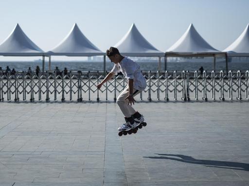 Ein junger Mann fährt Rollerblades. Im Hintergrund stehen Pavillons an einer Strandpromenade.