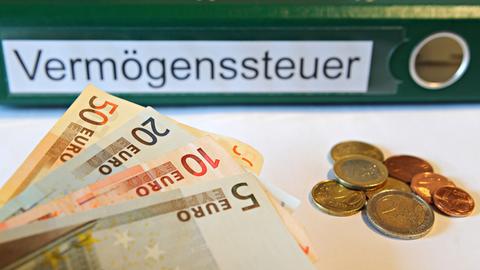 Ein Akten-Ordner mit der Aufschrift "Vermögenssteuer" liegt im Hintergrund. Davor sind Euro-Scheine und Münzen zu sehen.