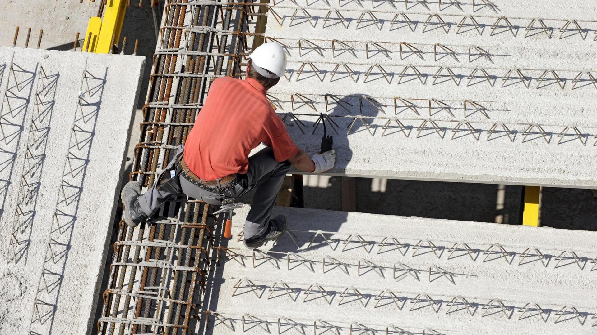 Ein Arbeiter trägt weißen Helm und klettert an einer Baustelle über die Stahlverstrebungen.
