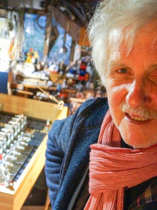 Ein Mann mit weißen Haaren und weißem Schnäuzer steht im Vordergrund des Bildes. Hinter ihm stehen lauter ungewöhnliche Geräte, Musikinstrumente, die er selbst gebaut hat.