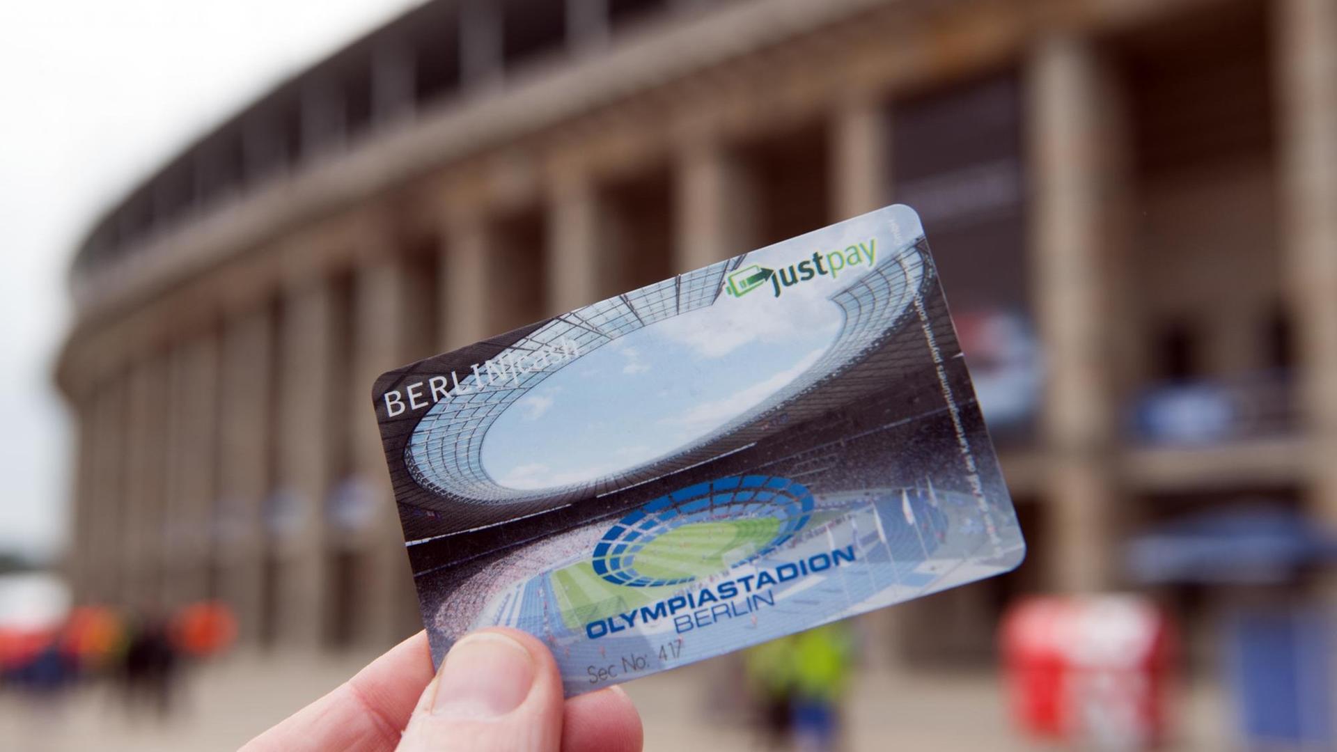 Die Hand eines Mannes hält eine Just-Pay-Karte vor das Berliner Olympiastadion. 
