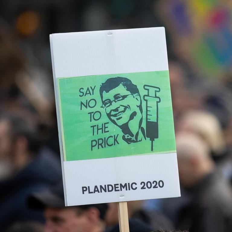 Bei einer Demonstration in England wird ein grünes Schild hochgehalten, auf dem der Kopf von Bill Gates und eine Spritze abgebildet sind.