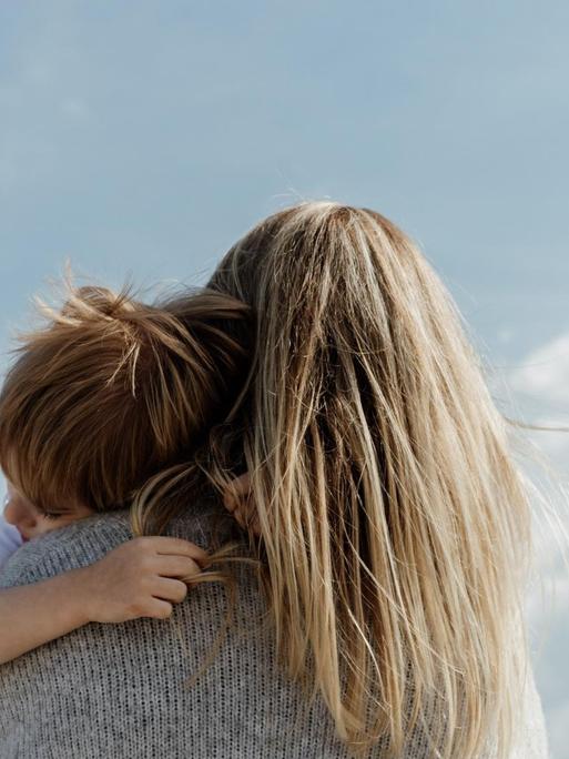 Eine Person mit langen blonden Haaren trägt ein Kind auf dem Arm. Sie sind von schräg unten aufgenommen, im Hintergrund erstreckt sich der blaue Himmel.