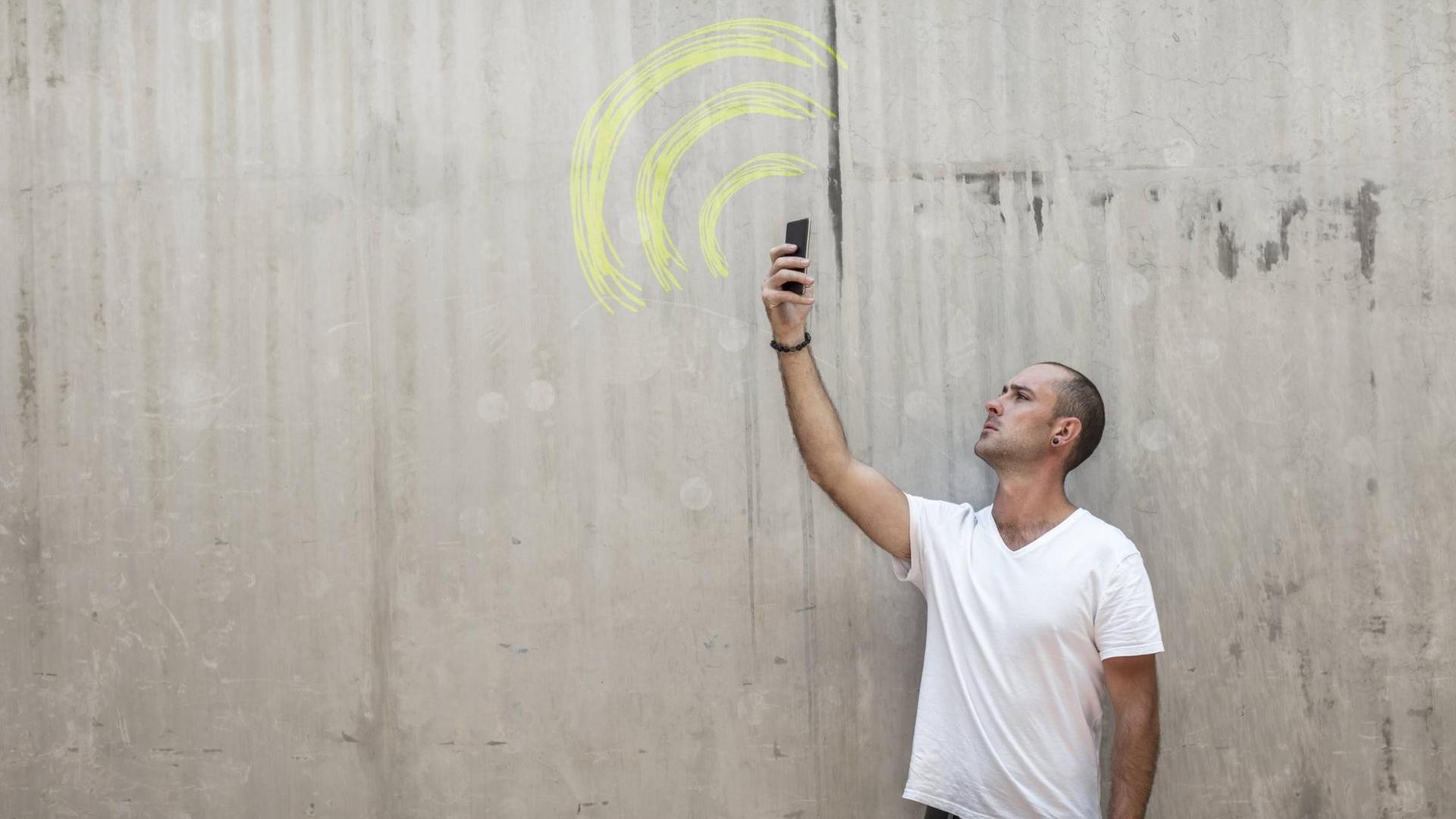 Mann hält sein Mobiltelefon in die Luft, auf einer Wand ist das WLAN Zeichen gemalt