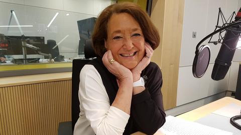 Eine ältere Dame sitzt in einem Radiostudio und lächelt in die Kamera, während sie den Kopf auf die Hände gestützt hat.