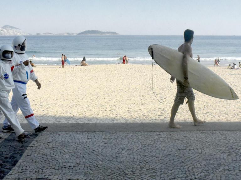 Ein Paar in Raumfahrer-Schutzanzügen flaniert am am Strand von Ipanema entlang.