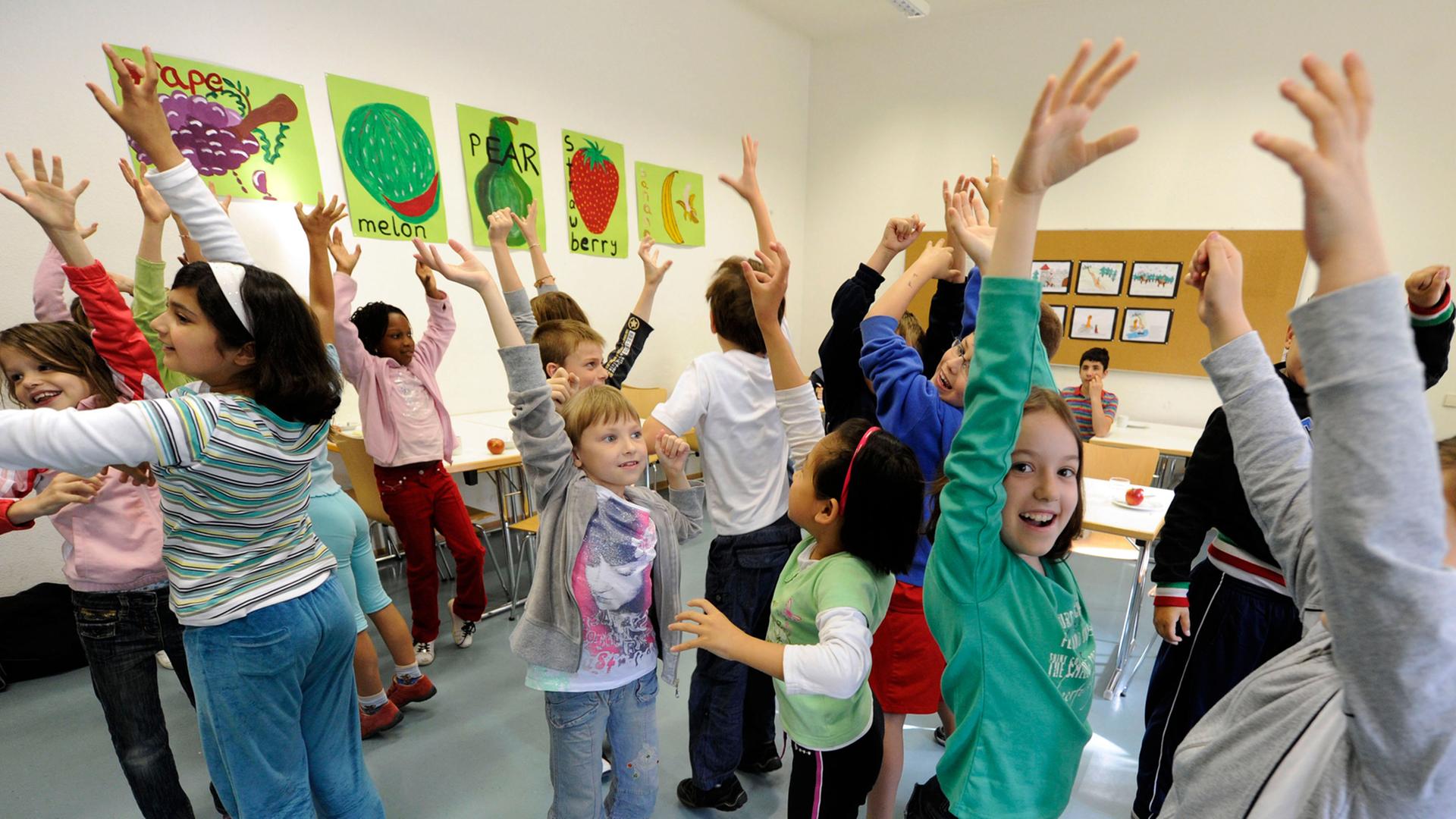Kinder stehen in einem Klassenraum und heben ihre Arme, an den Wänden bunte Bilder.