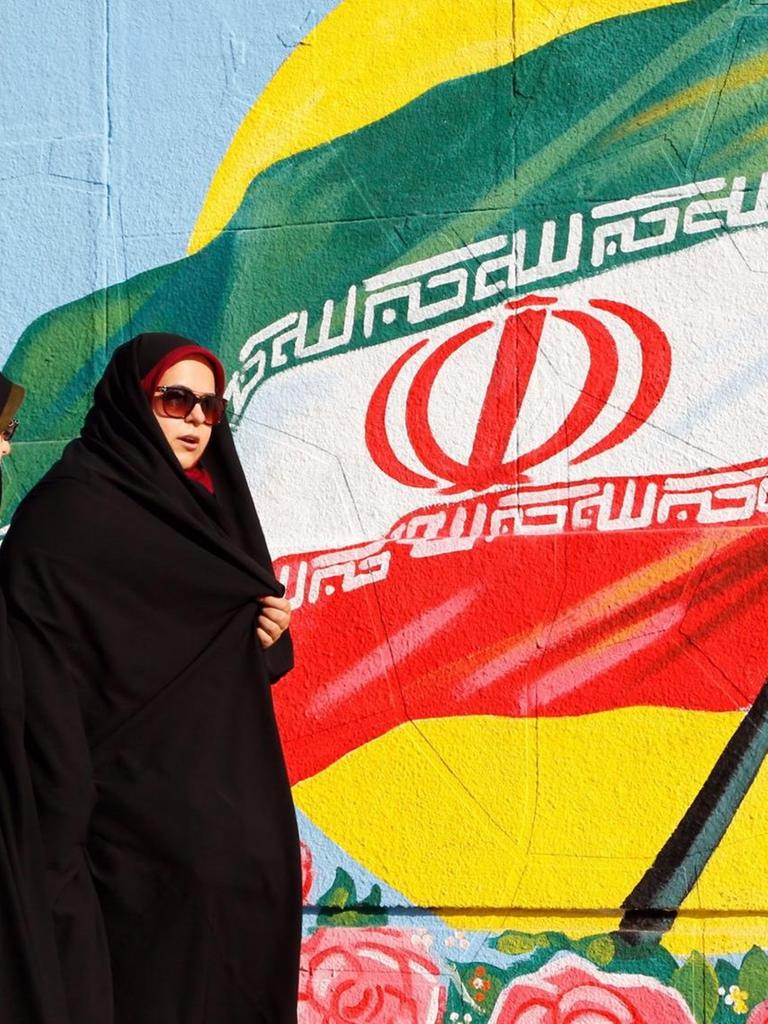 Zwei iranische Frauen in Teheran vor einer iranischen Flagge, die als Graffiti an eine Wand gesprüht ist