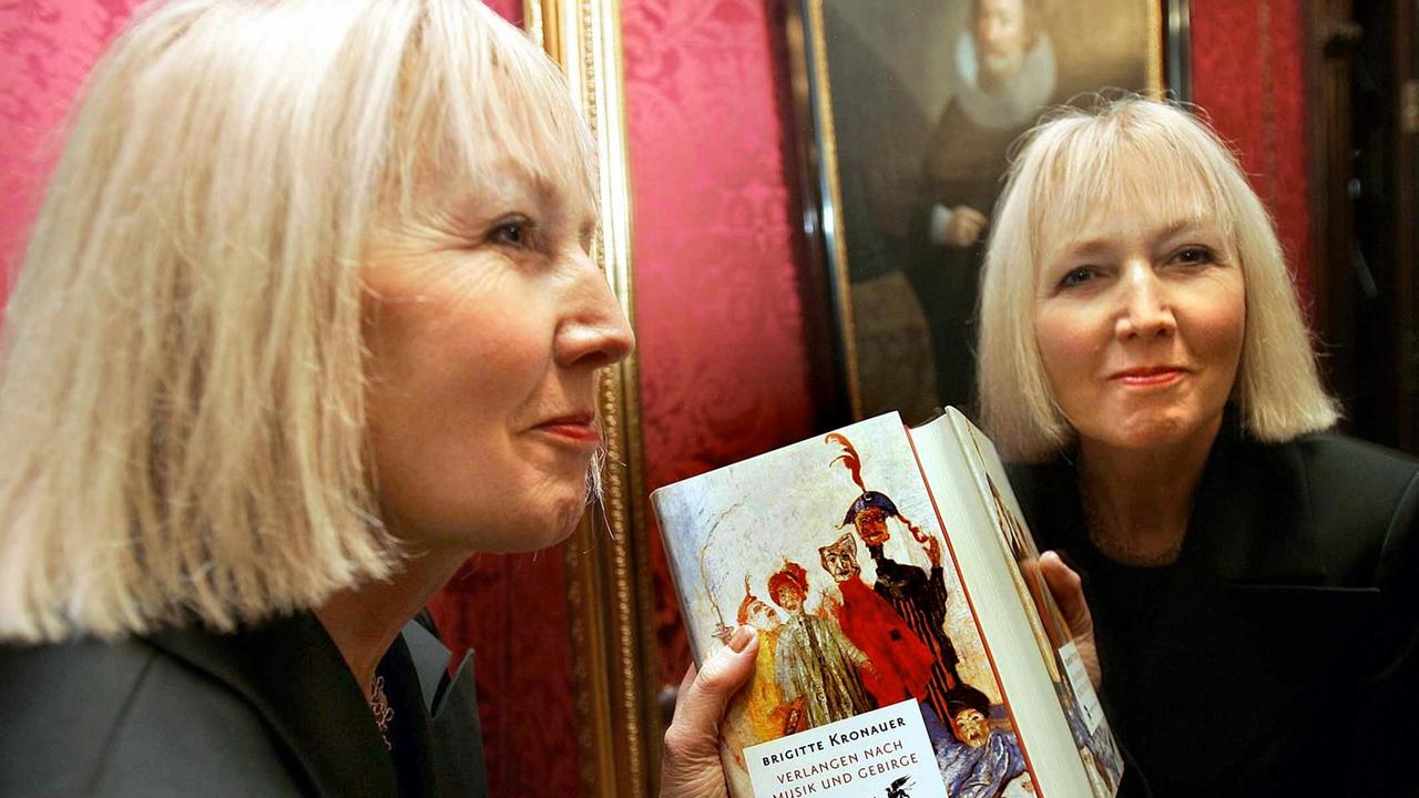 Die Schriftstellerin Brigitte Kronauer steht mit ihrem Buch vor einem Spiegel und blick uns aus diesem lächelnd entgegen. Sie erhielt 2005 in Darmstadt den Georg-Büchner-Preis.